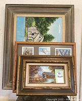 Three framed art pieces stacked: top small covered bridge painting, middle frame with three aviation prints, back large house by water painting.