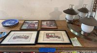 Photo of four framed art prints of vintage cars and farm children with a stack of four blue and white plates and a five-light metal hanging light fixture shown on a table.