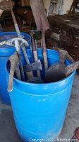 Blue plastic barrel container filled with various rusty garden tools with wooden handles including shovels, ax, and more.
