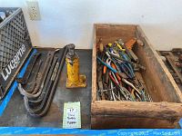 Close up image of various steel pry bars and crowbars stacked together with visible wear and rust, beside the yellow hydraulic jack