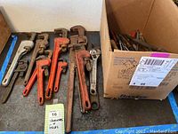 Wide view showing pipe wrenches arranged on a surface beside a cardboard box containing various files and screwdrivers.