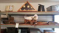 Photo showing military folded flag in wooden glass display case, miniature white canvas-covered wooden stagecoach, black leather holster, metal pitcher, and wash tub on metal shelving.