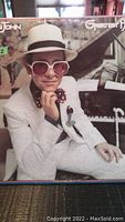 Front cover of the 'Elton John Greatest Hits' vinyl record album showing Elton John seated at piano in white suit, hat, and large pink glasses.