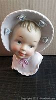 Front view of porcelain vintage baby face head vase with bonnet hat decorated with white roses and pink bow detail on collar.