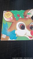 Front cover of the vintage shaped book showing the title The Rudolph the Red-Nosed Reindeer Book with holly and a blue bow, featuring Rudolph's face
