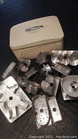 Assortment of vintage metal cookie cutters alongside cream-colored old tin box with metal handle, showing variety of shapes and sizes