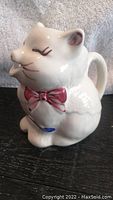 Front and side view of white ceramic cat-shaped creamer with red bow tie and closed eyes detail.