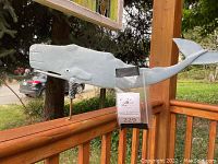 Blue-gray ceramic whale mounted on stand attached to railing outside porch on wood deck, visible car and trees in the background.
