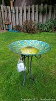 Outdoor shot of birdbath showing blue and gold iridescent glass basin with peacock feather design on a black metal stand, placed on grass near a wooden fence.