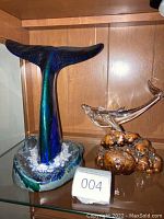 Two whale sculptures on glass shelf inside wooden cabinet: ceramic blue whale tail sculpture and clear glass whale on varnished wood base.