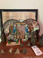 Overview of entire lot showing framed papyrus, two metal plates, colorful Egyptian-themed picture frame with scarab beetle, four Canopic jars, and six pyramids paperweights arranged on a wooden surface.