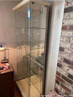 Full frontal view of the tall glass display cabinet showing transparent glass panels and four shelves inside with a cream-colored base.