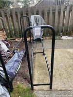 Full side view of the black metal tubular outdoor firewood rack on a concrete patio next to a wooden fence. A grey protective cover is draped over one end of the rack. Ground around rack includes grass and paved surfaces.