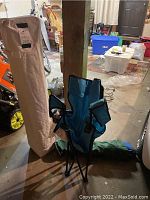 Beige carrying bag for pop up canopy standing upright next to teal folding chair in carrying bag in garage setting.