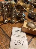 Overall view of silver and silver-plated items including soldiers salt and pepper shakers, gravy bowl, small bowls, and a boxed spoon with red ribbon.