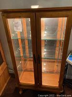 Full front view of light-finish curio cabinet with two glass doors and internal glass shelves, interior lighting illuminated