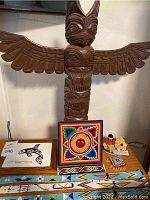 Front view of the wooden totem pole with outstretched wings alongside a colorful square beaded patch, beaded belt, and beaded necklace and braces displayed on a wooden surface.