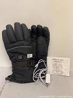 Photo of pair of black heated gloves standing upright, showing power button on one glove, adjustable wrist straps, included white cord charger and user manual beside gloves.