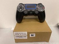 Black PlayStation 4 DualShock 4 controller resting on a cardboard box with a label, showing front view with buttons, touchpad, and analog sticks visible.