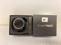Black smart watch inside an open black box showing the watch face and strap. Box lid displaying 'Smart Watch' text.