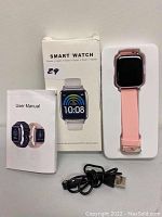 Photo showing pink smart watch in white packaging, user manual booklet, and USB charging cable on white surface.