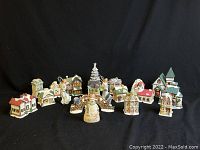 Wide shot of the full set showing 18 distinct Christmas village figurines arranged in a row on a black background. Includes houses, churches, snowman, and other holiday figures.