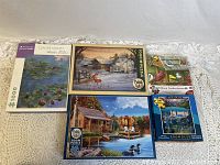 Five assorted jigsaw puzzles on lace fabric. Two 1000-piece puzzles include Claude Monet's Water Lilies and a winter rural scene by Cobble Hill. Three 500-piece puzzles include a birds collage, a lake with ducks and cabin by Dowdle, and a Lake Louise one.