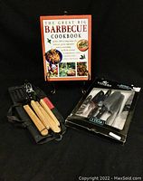 Wide view of all items in the barbecue lot including The Great Big Barbecue Cookbook standing on a black stand, four-piece barbecue tool set laid on a black pouch, and Winer Spectrum knife set in original packaging.