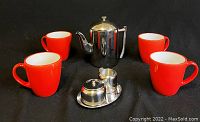 Four orange ceramic mugs and a polished metal teapot with strainer on a black background. Also includes metal sugar and creamer containers on a small tray.