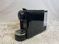 Black Martello Stilista Primeo espresso capsule machine, shown on lace fabric background. Side profile with water reservoir visible.