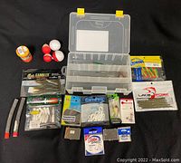 Photo showing fishing tackle box open with assortment of fishing lures, hooks, bobbers, fishing line, and other equipment spread around it.