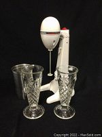 Front view of white Hamilton Beach Drink Master milkshake machine with two tall diamond-patterned glasses and stainless steel mixing cup on black background.