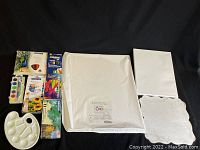 Overview photo of multiple art supplies including acrylic paints sets, paint palette, digital art paper, and blank canvases.