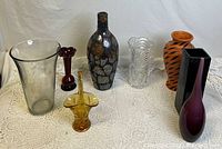 Photo of eight assorted vases and glass décor pieces on white lace tablecloth, demonstrating variety of styles, colors, and sizes ranging approx 9-16 inches tall.