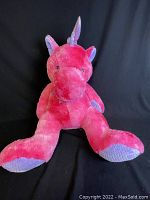 Photo shows front-facing pink plush unicorn against a black background. The unicorn is seated with legs extended forward and arms to the sides. The horn, ears, wings, and feet bottoms have lavender sparkle fabric accents. The unicorn's eyes are black sewn or button style. The plush has a large snout and round body shape.