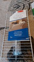 Photo of Rubbermaid 61 cm Linen Shelf Kit packaging placed on wire shelf showing product information and accessories included.