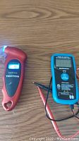 Mastercraft battery tester shown with test leads on a wooden surface alongside Tekton electronic tire pressure gauge with a lit display and LED light