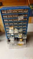 Blue metal organizer with 48 small drawers and 4 larger bottom drawers containing various metal hardware parts. Front view showing many drawers filled with screws, nails, bolts and other items.