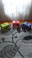 Three folding plastic stools in lime green, red, and blue colors arranged on a patterned carpet in front of a sheer curtained window.