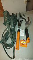 Photo showing green extension plug with multiple outlets, hand rake with orange handle, and hand claw with black and orange handle on a table
