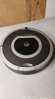 Front perspective photo of gray and black iRobot Roomba robotic vacuum cleaner on flat surface, showing circular shape and top controls.