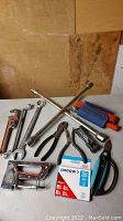 Photo showing multiple hand tools including wrenches, pliers, utility knives, scissors, a staple gun, and a box of staples on a white table with a wood background.