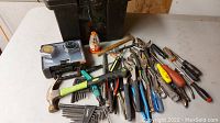 Mixed hand tools including hammers, pliers, screwdrivers, hex keys, and carpenter's level arranged on a surface in front of a black toolbox.