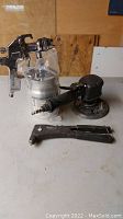 Photo of three tools on a workbench: a metal paint spray gun with a metal paint cup, a black pneumatic air sander, and a metal staple gun. The tools show signs of use and wear.
