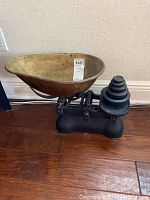 Antique brass and iron scale showing the large brass weighing pan and iron base with beam balance, positioned on a wooden floor.