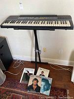 Full view of Casio CTK-480 keyboard on X-style stand, with music books laid in front on floor.