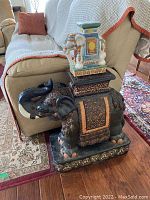 Photo shows large black ceramic elephant garden bench with detailed gold designs and a smaller colorful ceramic elephant figurine on top next to a couch.