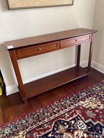 Wooden console table with two drawers and a bottom shelf; viewed from an angle showing the right side and front; placed on a hardwood floor next to patterned rug.