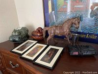 Side view of ceramic hippo figurine, ram-shaped basket, leather horse figurine, framed batik prints and electronic device on a wooden surface.