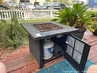 Fire pit shown outdoors on a patio, black square unit with propane tank in storage compartment below.